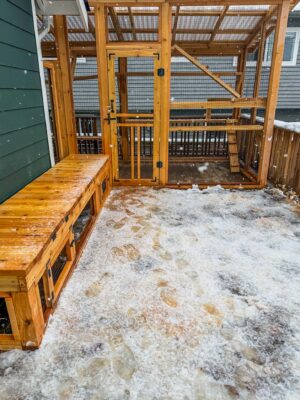 Custom-built wooden catio attached to a Seattle home during light snowfall, featuring a secure mesh enclosure, wooden framing, a hinged entry door with latch, interior climbing ramp and elevated shelving, and a built-in bench with storage along the exterior; snow-covered ground with visible footprints leads up to the enclosure.