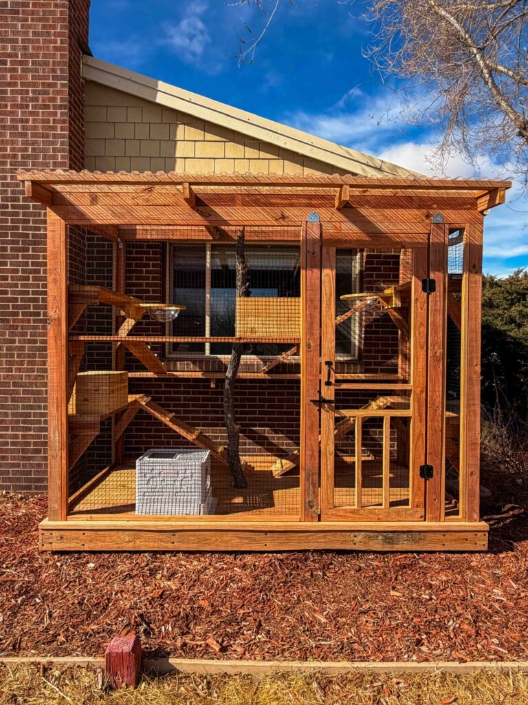 Exterior view of a custom cedar catio enclosure attached to a brick home in Ketchum, Idaho featuring wire mesh panels, a secure entry door, a sloped transparent roof, multiple interior climbing shelves and ramps, a natural tree trunk climbing post, and an integrated window entry for cats.