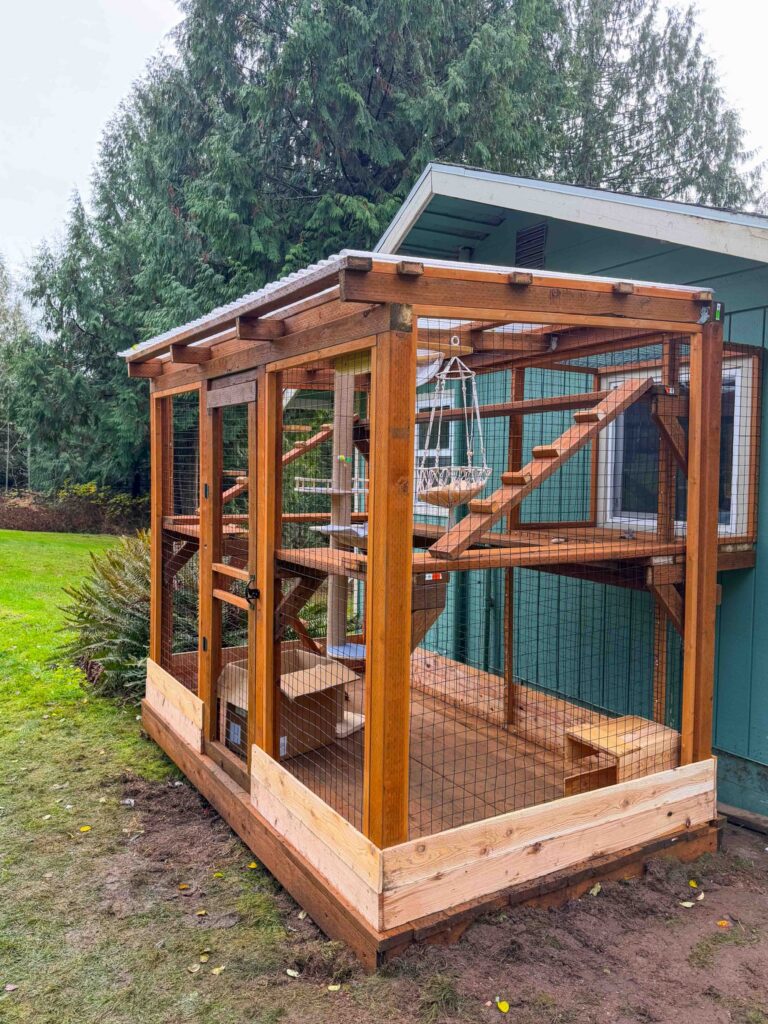 Large custom wooden catio enclosure attached to a home in Sherwood, Oregon featuring elevated platforms, cedar climbing ramps, a hanging macramé cat hammock, secure wire mesh panels, and a clear roof covering a spacious two-level outdoor cat enclosure surrounded by trees and backyard landscaping.