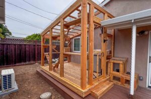 Large custom-built wooden catio attached to a home with wire mesh walls, clear polycarbonate roof, multiple levels, ramps, and a secure entry door on a raised deck platform.