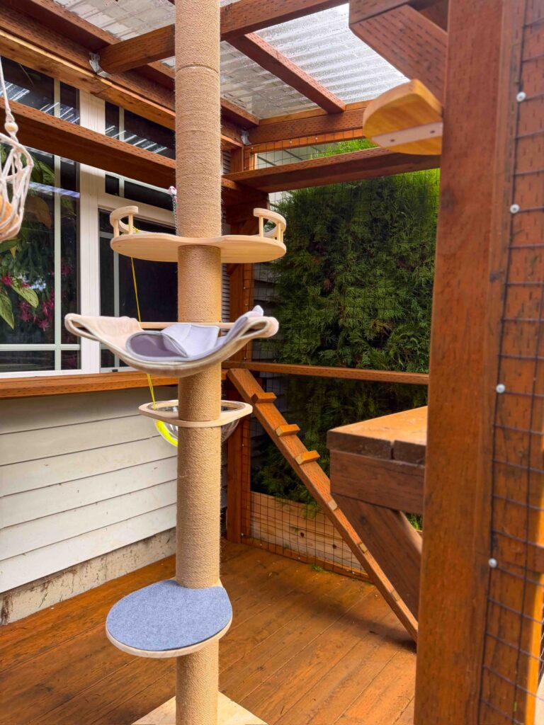 Interior view of a custom wooden catio attached to a home in Sherwood, Oregon, featuring a tall floor-to-ceiling sisal scratching post cat tower with multiple platforms, a hammock-style cat bed, hanging enrichment toys, cedar climbing ramps, and enclosed wire mesh walls beneath a clear corrugated roof that allows natural light to fill the spacious outdoor cat enclosure.