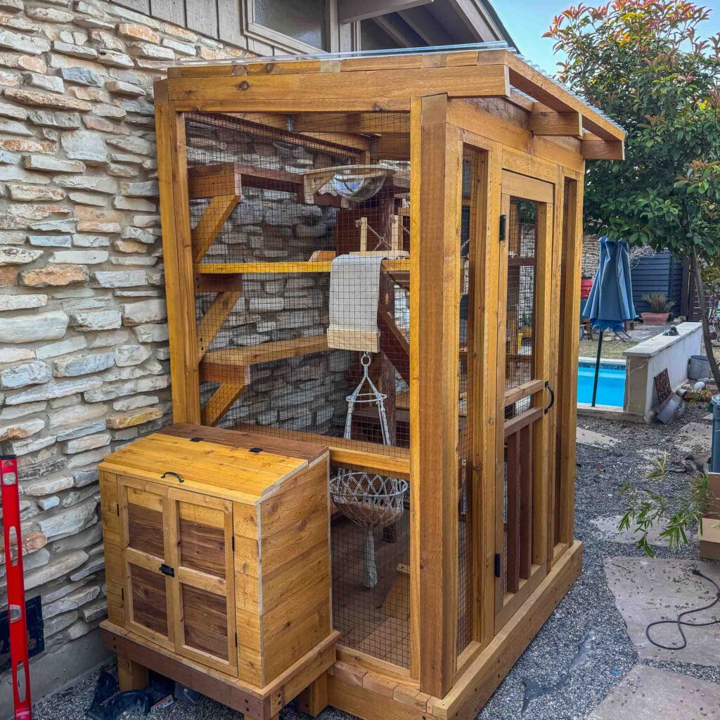 Compact custom catio enclosure with a wooden storage box attached, featuring multiple interior platforms, climbing ramps, and a visible hanging hammock inside