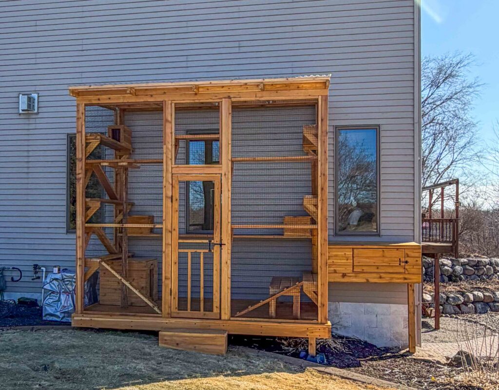 Exterior view of a custom freestanding wooden catio attached to a home in Sunfish Lake, MN with full-height wire mesh panels, secure entry door, multiple interior climbing shelves, and a connected elevated tunnel leading from a house window.
