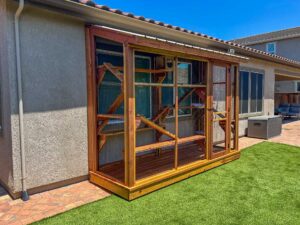 Long rectangular wooden catio enclosure with mesh panels and built-in ramps and platforms, installed along the exterior wall of a home overlooking a landscaped backyard