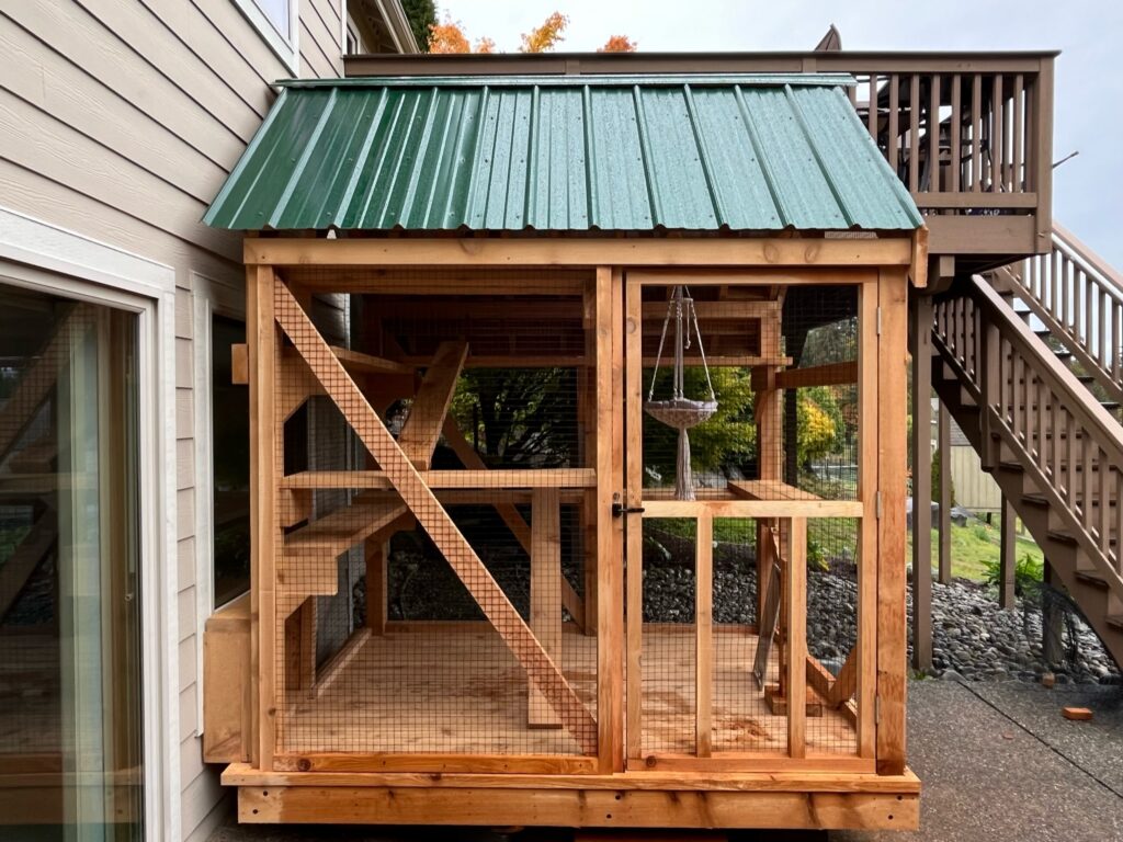 Exterior view of a custom-built wooden catio in Woodland, MN with a green sloped metal roof, enclosed wire mesh walls, a full-height access door, and interior climbing structures attached to the side of a house near an elevated deck and staircase.