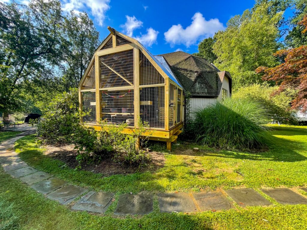 Exterior view of a large custom wooden catio attached to a home in Stockton, NJ, with a barn-style roofline, galvanized wire mesh panels, elevated foundation, and landscaped garden surroundings under a bright blue sky.