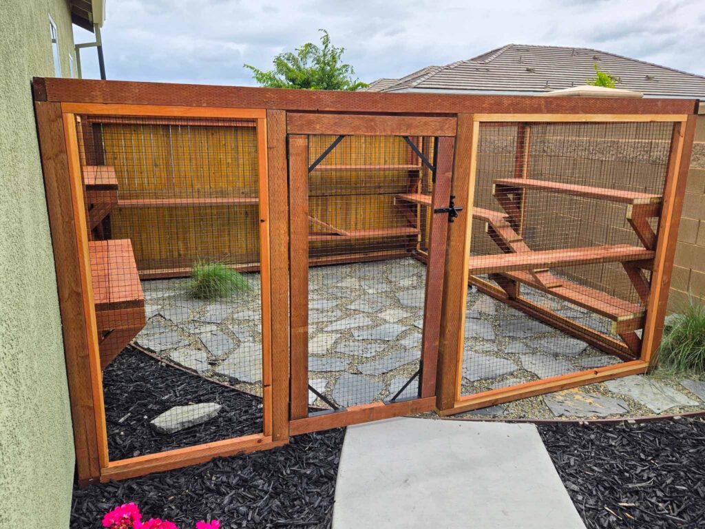 Front view of a custom outdoor catio in Galt, CA with a centered wooden entry gate reinforced with black metal brackets, enclosed by wire mesh panels, and featuring multi-level wooden shelves and ramps over a stone and gravel floor with landscaped garden beds nearby.