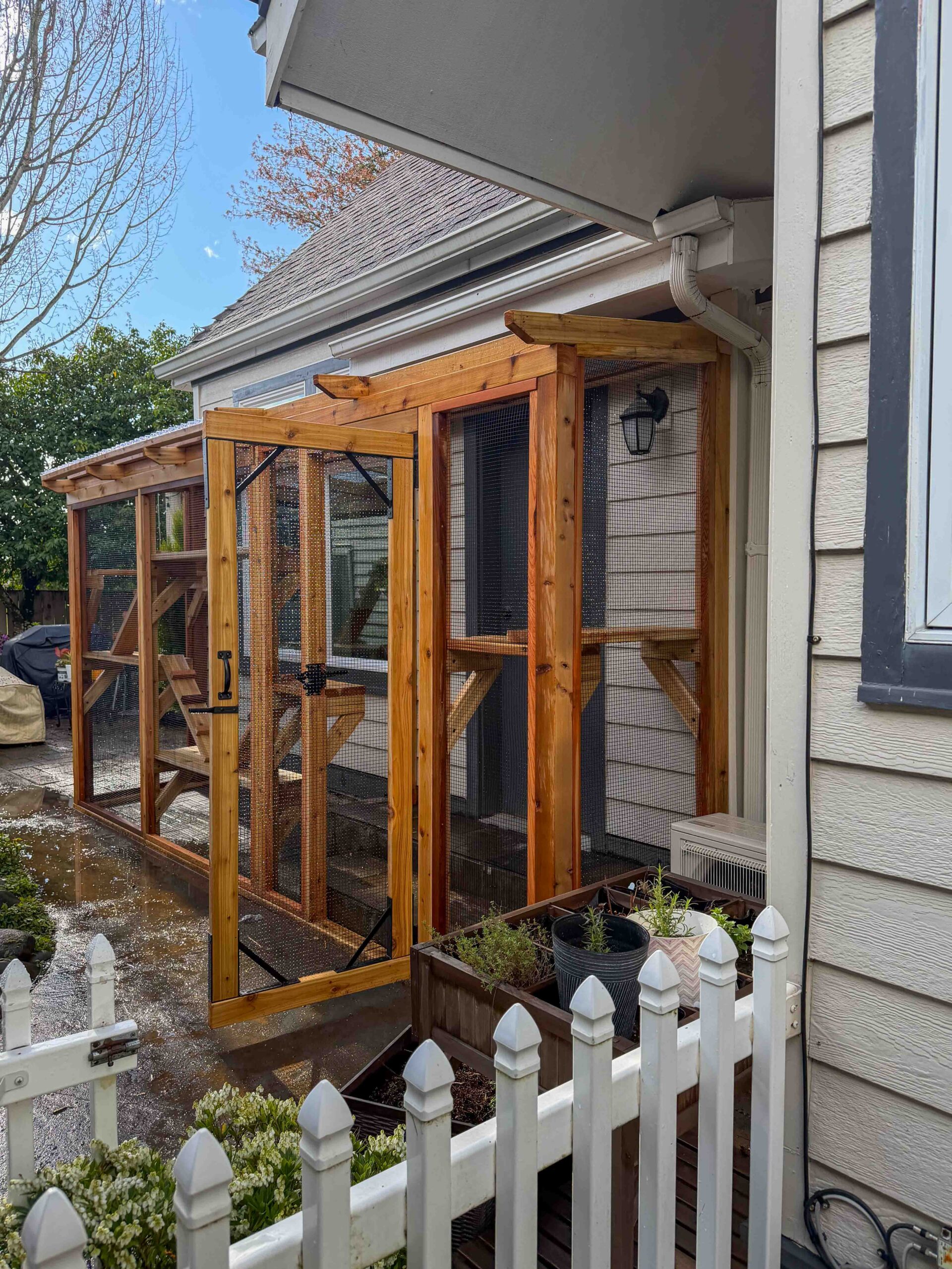 Angled view of a custom catio with an open mesh door, showing wooden framing, interior platforms, and placement next to a small patio and garden area.