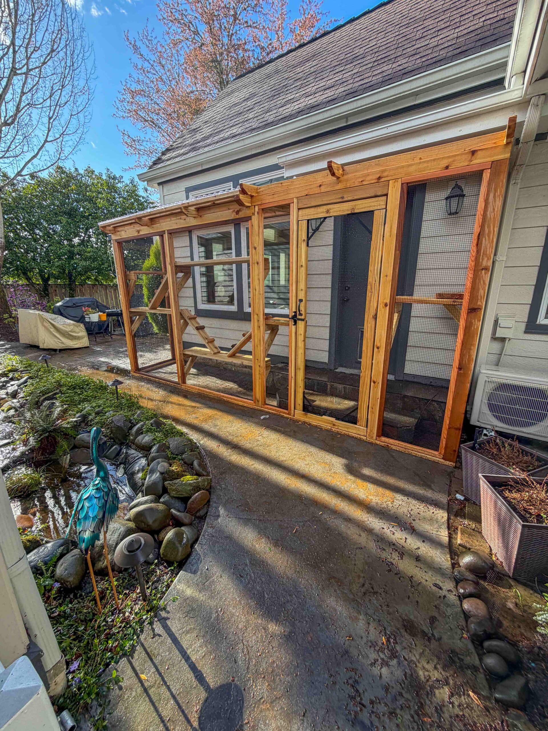 Custom-built wooden catio attached to the back of a home featuring a long enclosed structure with wire mesh walls, a clear roof panel, and multiple interior climbing platforms beside a landscaped garden with rocks and a small water feature.