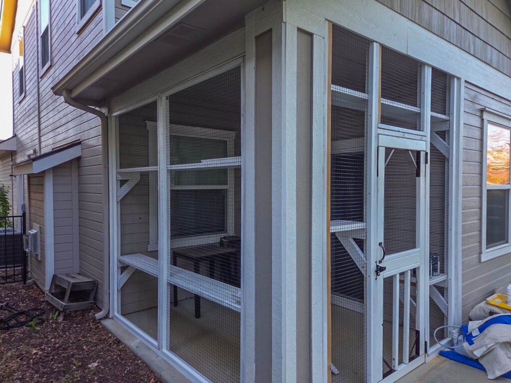 Custom screened catio attached to a two-story home in Boise, Idaho, featuring white framing, secure wire mesh panels, multiple wall-mounted cat shelves, elevated lounging platforms, and a covered roof design for year-round outdoor enrichment.